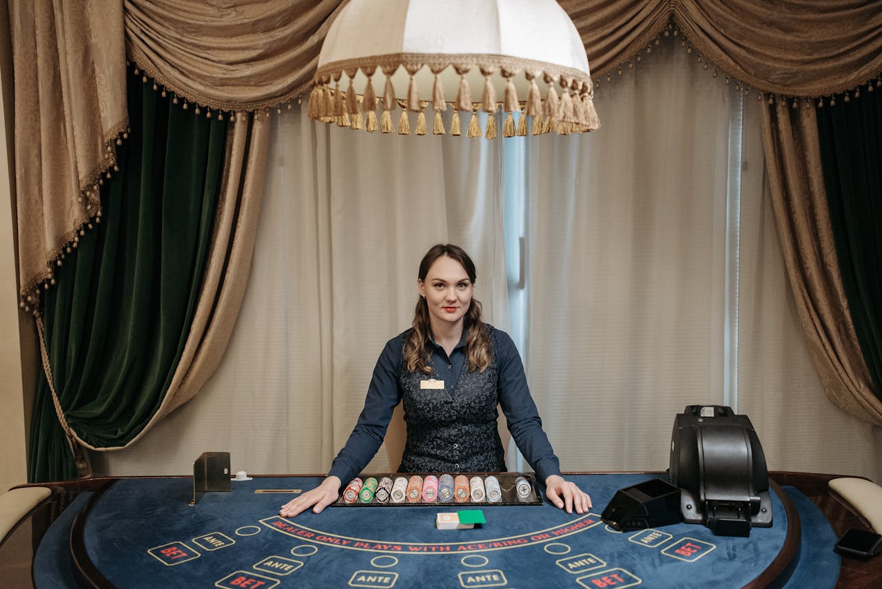 A smiling female casino dealer at a gaming table surrounded by chips and cards indoors.